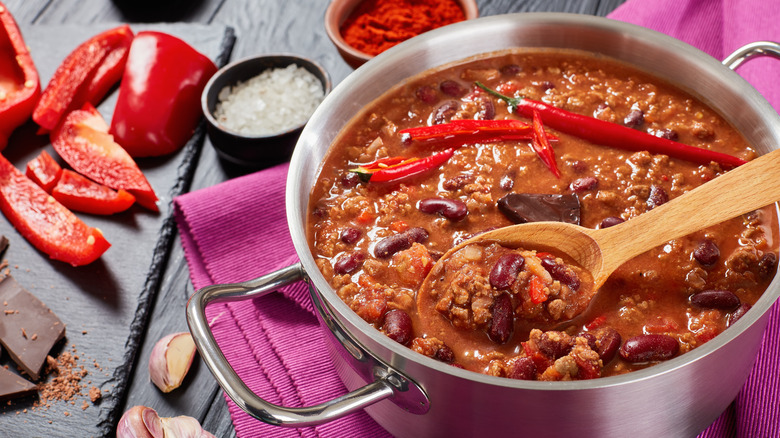 Pot of chili with peppers and seasonings on table with wooden spoon