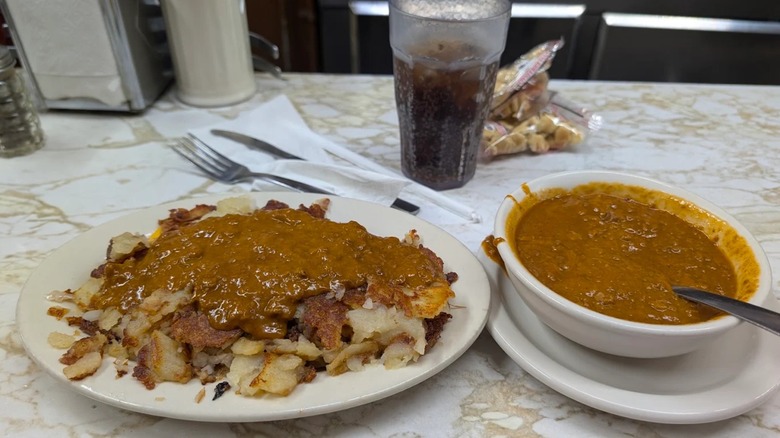 Bowl of chili on counter with chili on home fries and glass of soda