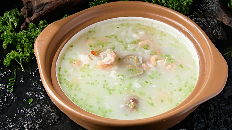 Shrimp congee in a clay pot on a black table surrounded by parsley and salt