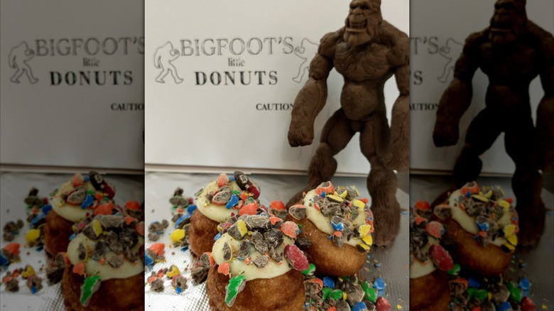 Close up of donuts topped with crushed candy in front of a sign for Bigfoot's Little Donuts and a Bigfoot figurine