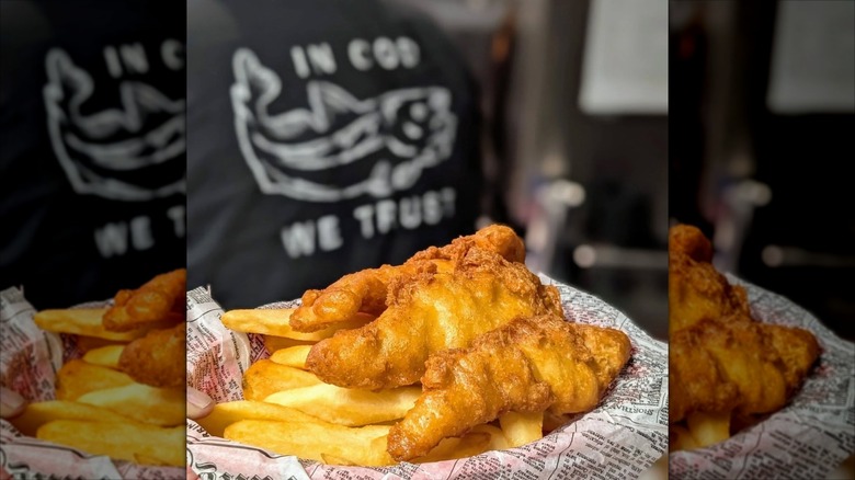 Basket of fish and chips on newspaper with blurred shirt in background reading "In Cod We Trust"