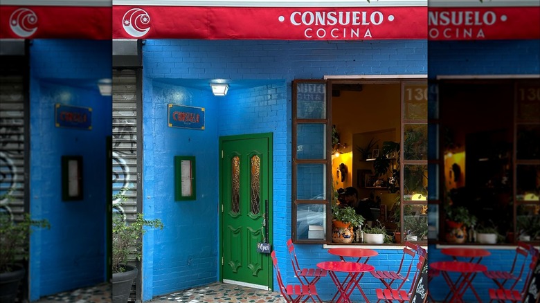 The painted blue brick exterior of Cocina Consuelo with a red awning and red table and chairs outside