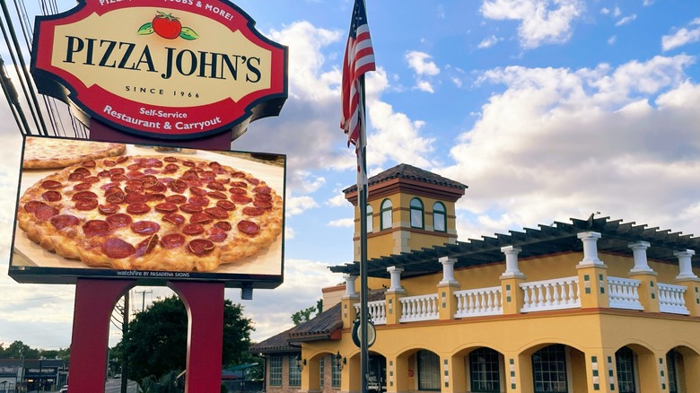 The yellow walled exterior of Pizza John's with a sign showing a pepperoni pizza and an American flag outside