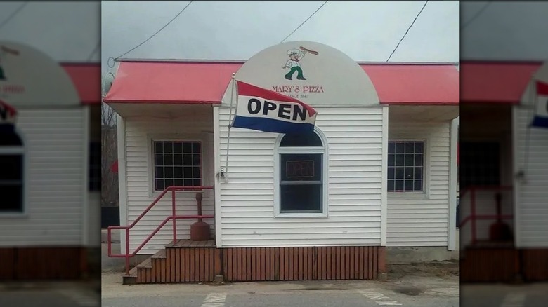 The white clapboard exterior of Mary's Pizza with a blue, red, and white flag outside that says 'open'