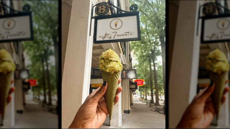 Hand holding an ice cream cone with yellow ice cream below the exterior sign for eighty7sweets
