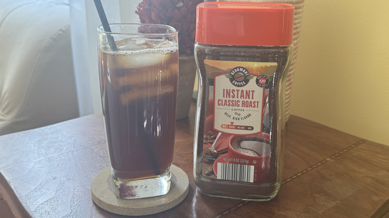 A glass of iced coffee on a table sitting next to a cannister of Beaumount Instant Classic Roast