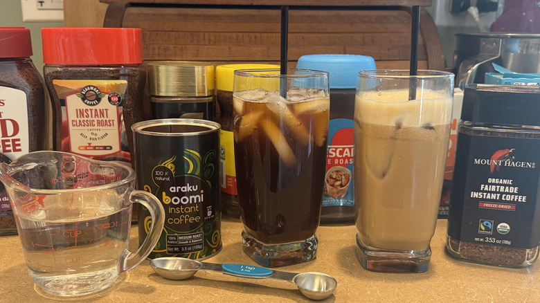 An assortment of instant coffees, glasses, and measuing utensils on a kitchen cabinet