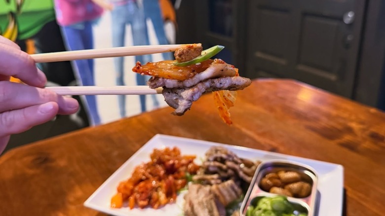 hand holding meat and vegetables with chopsticks over blurred plate on table