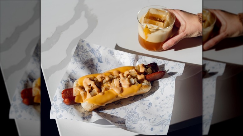 Loaded hot dog on paper with hand holding cocktail glass