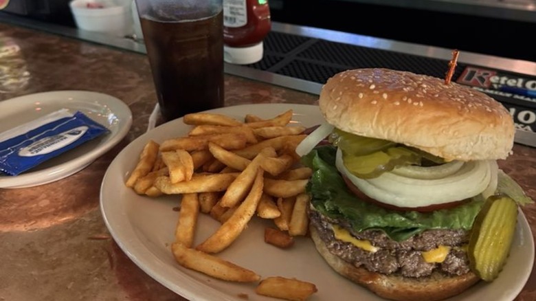 Tall cheeseburger with onions and pickles on plate with fries and drink on bar