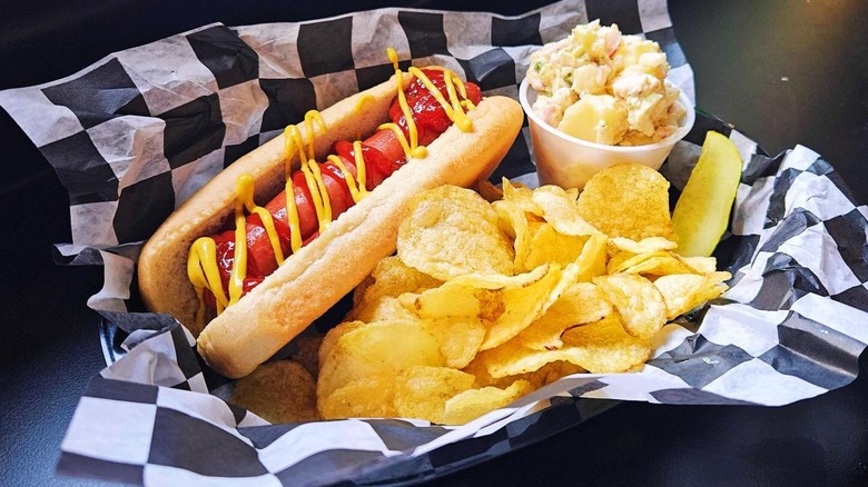 Basket with black and white, checkered paper, hot dog, potato chips, and container of potato salad