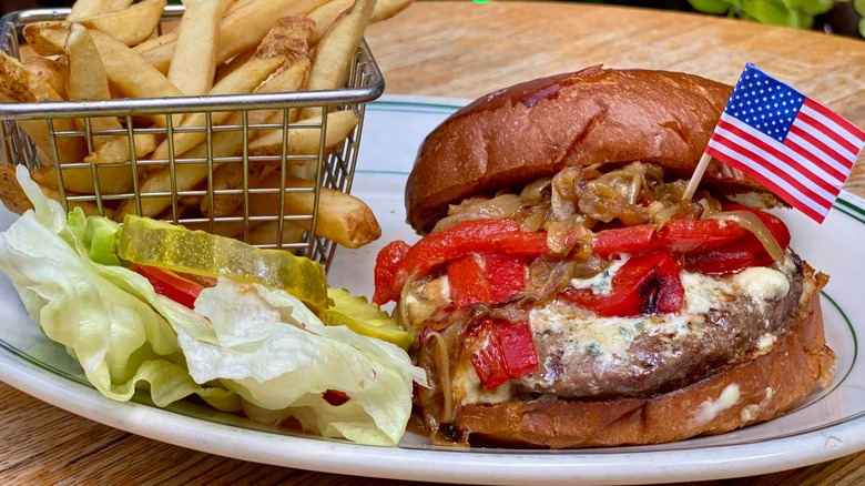Burger with onions and peppers on plate with basket of fries, lettuce, and pickle