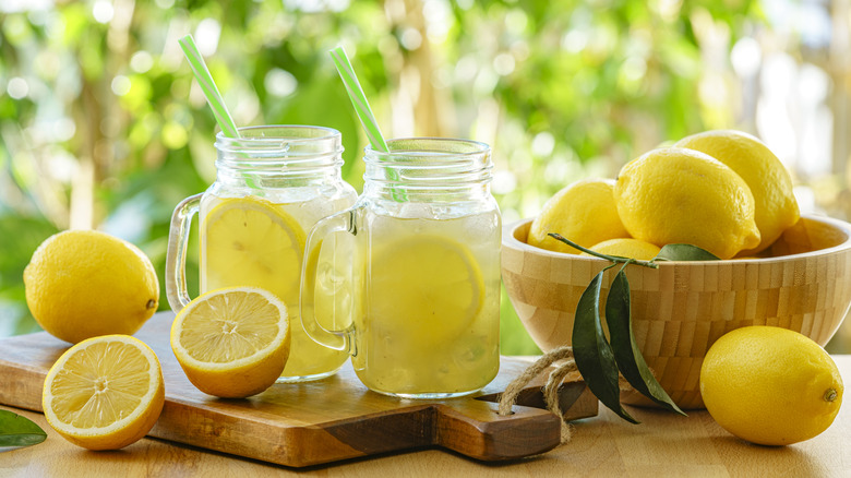 Mason jar mugs of lemonade are styled with a bowl of lemons and sliced lemons on a cutting board.