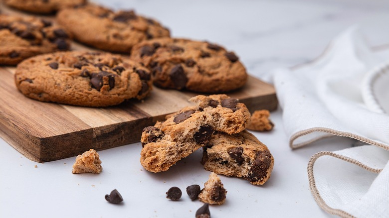 A broken chocolate chip cookie in front of a wooden platter full of whole chocolate chip cookies