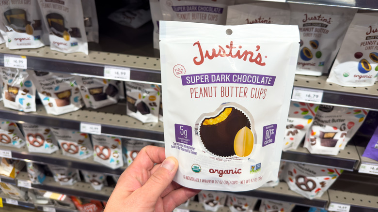A person's hand is seen holding a package of Justin's peanut butter cups in a store aisle.
