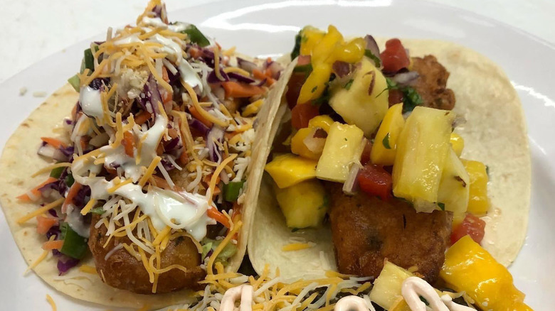 A generous plate of fish tacos. The one on the left is sprinkled with ginger coleslaw, and the one on the right is topped with mango salsa.