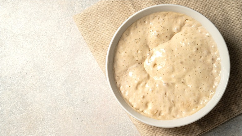 Top-down view of bubbly sourdough starter proofing in a white bowl over a tan cloth napkin