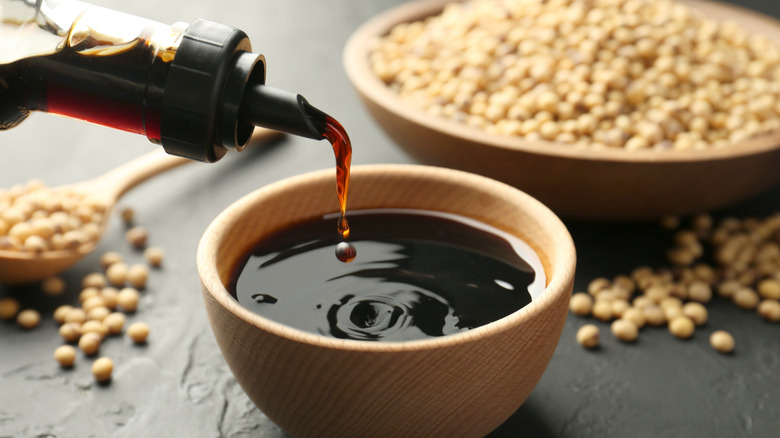 Soy sauce being poured into a small wooden bowl with soybeans in the background