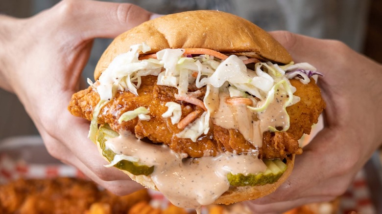Hands hold fried chicken sandwich with coleslaw and sauce.