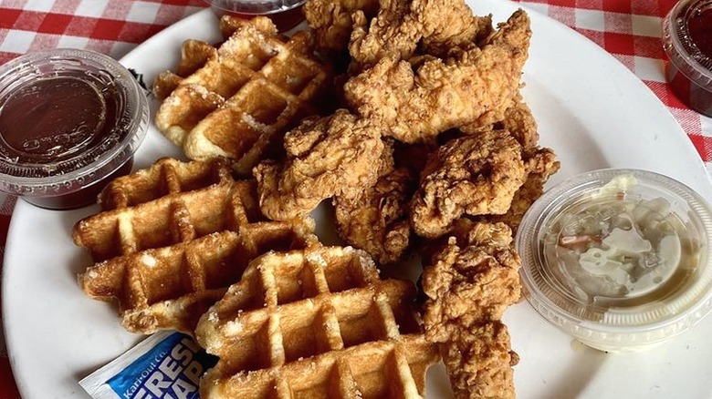 Close-up of a white plate with fried chicken, waffles, and plastic containers with syrup and sides.