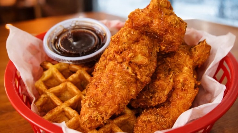 Pieces of fried chicken in red basket with waffle and sauce.