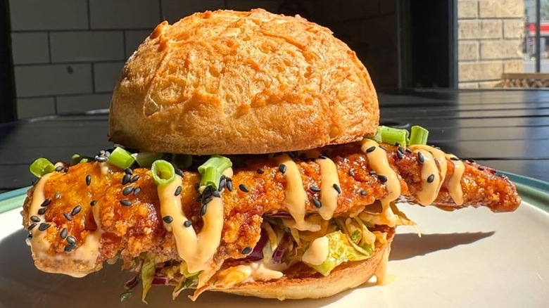 Massive piece of fried chicken in sandwich with lettuce, sauce, black sesame seeds, and scallions.