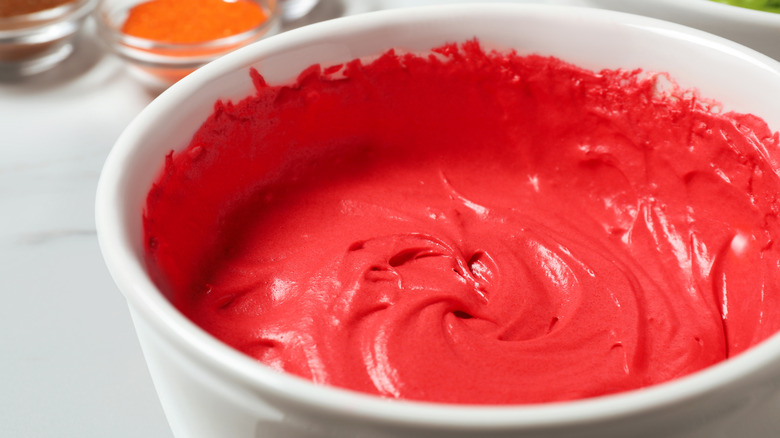 A white bowl full of red frosting with coloring agents in smaller bowls behind it