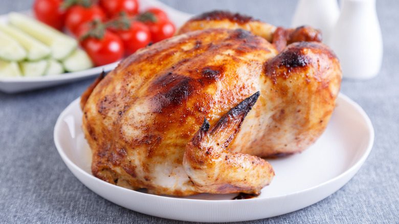 A rotisserie chicken is on a white plate with fresh vegetables in the background.