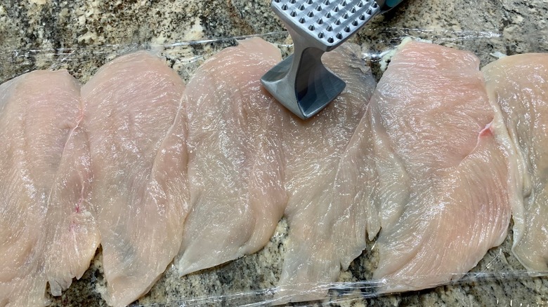 A cook using a meat mallet on a piece of meat covered with plastic wrap