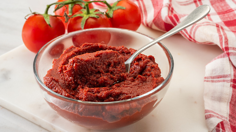 A bowl of tomato paste with whole tomatoes on the vine behind it and a quaint dish towel on the side.