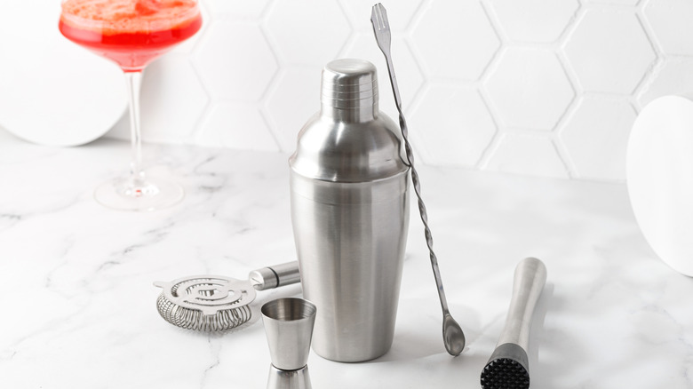 Stainless steel cocktail tools sit on a white countertop in front of a red cocktail