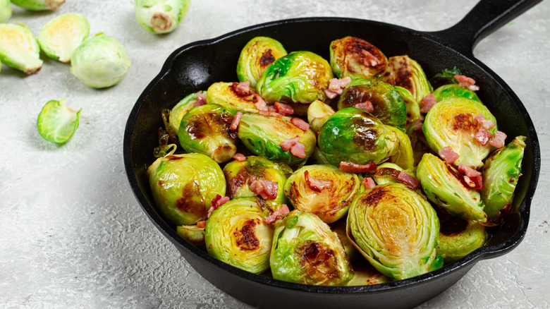 Caramelized roasted Brussels sprouts in a cast iron pan, topped with bacon, surrounded by raw Brussels sprouts on a white countertop.