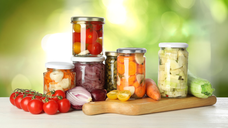 Jars of pickled vegetables on a white countertop and cutting board: carrots, cauliflower, tomatoes, zucchini, with fresh vegetables in front.