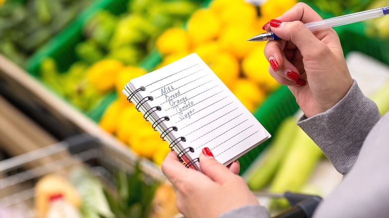 Food shopper with red-painted nails holding a grocery list in the produce section of a supermarket