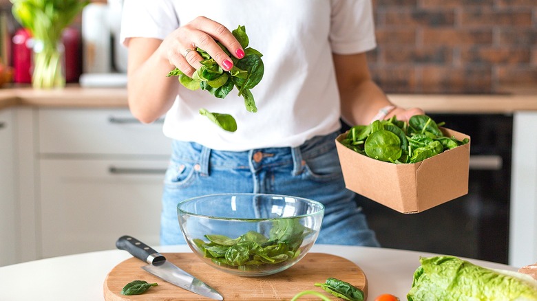 Person in a white t-shirt and jeans putting fresh spinach in a glass bowl over a knife and cutting board.