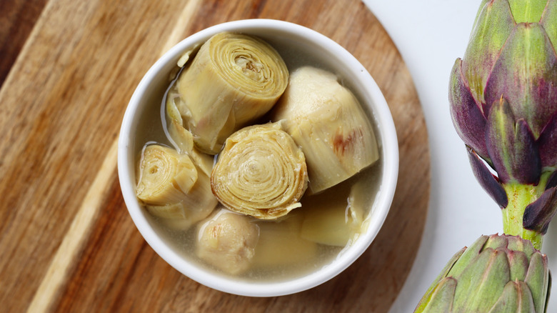 Brined artichoke hearts in a white bowl on a wooden board next to whole raw artichokes