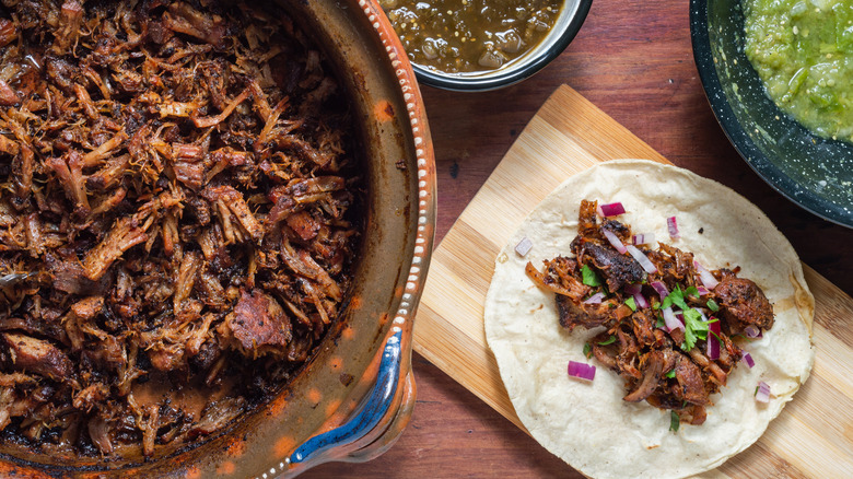 Serving dish of pork carnitas next to a taco and two sauces