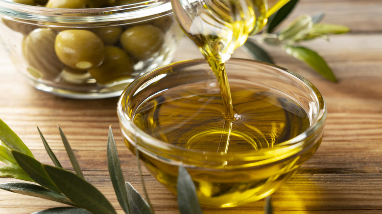 olive oil being poured into a glass bowl, with a bowl of fresh olives in the background