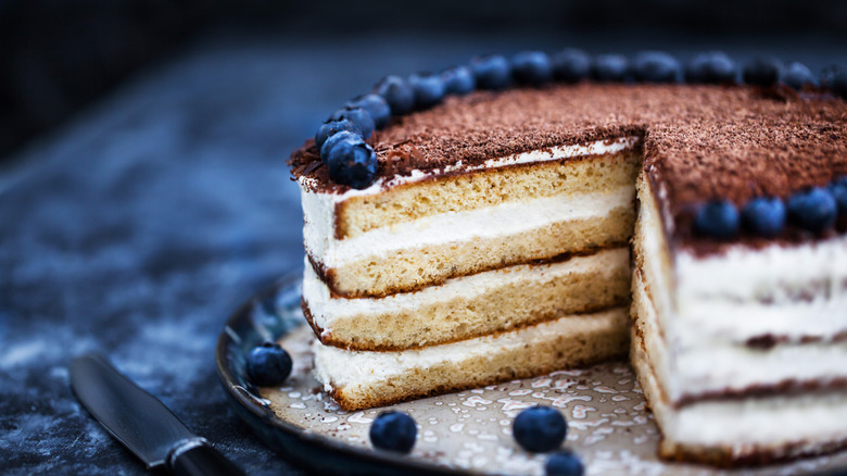 A four-layer cake decorated with frosting and blueberries with a slice already removed
