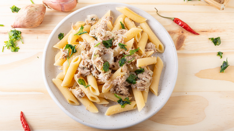 White bowl of pasta penne with canned tuna fish and herbs on wood surface.