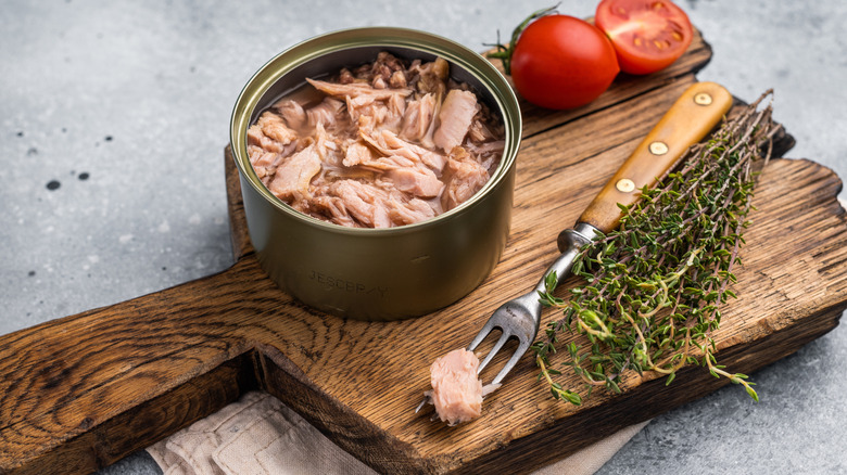Open can of tuna with tomatoes and herbs on wooden board.