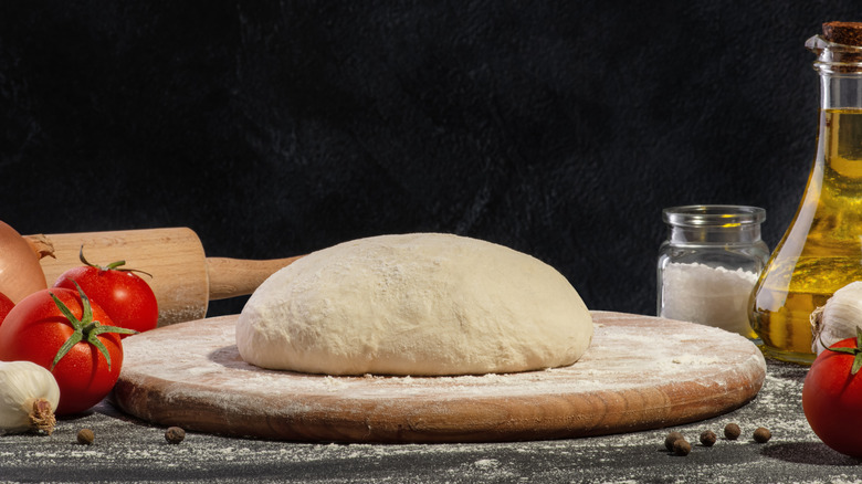 A dome of pizza dough rests on a floured wooden board; other ingredients like tomatoes and oil are also arranged in the frame.