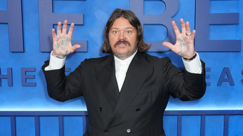 Actor Matty Matheson poses with his hands up in front of a blue wall.