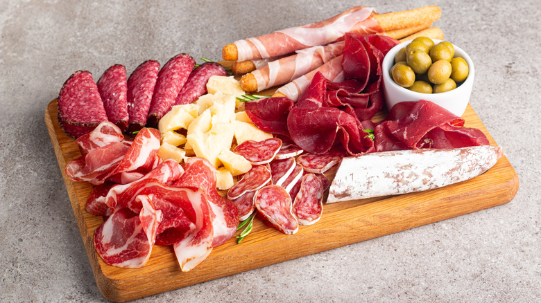 A charcuterie board containing meat, cheese, green olives, and breadsticks.