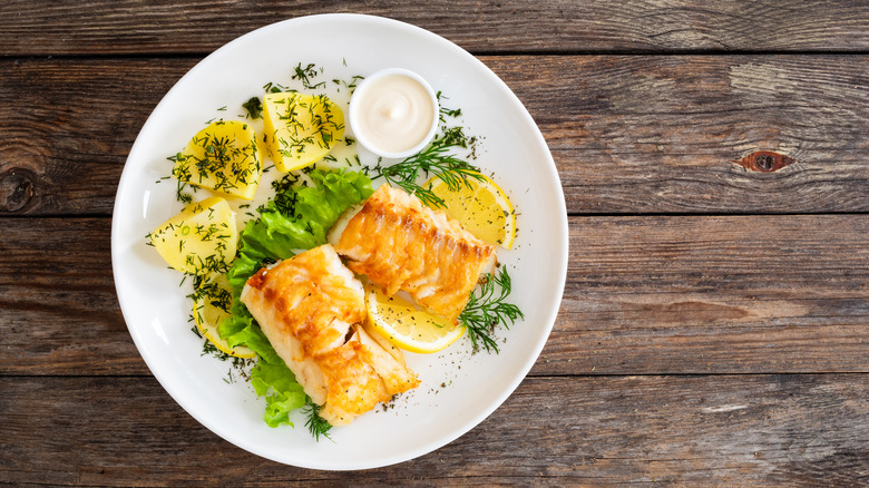 Grilled white fish with greens and lemon on a white plate atop a wooden table