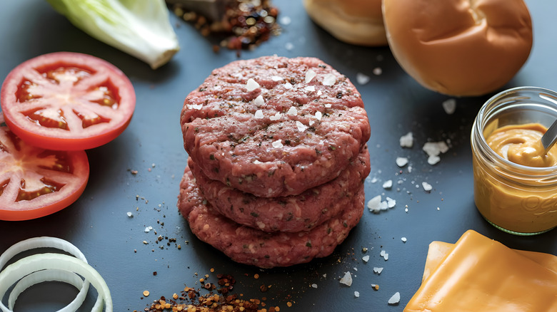 Traditional toppings like raw onions, sliced tomatoes, and cheese are arranged a stack of uncooked burger patties.