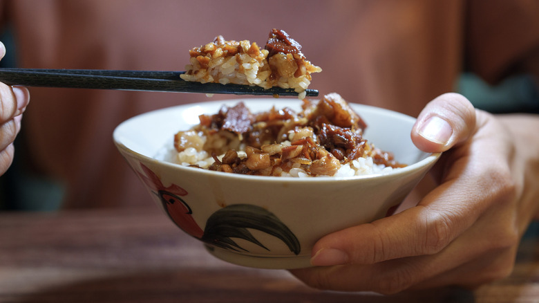 Hand holding chopsticks with Chinese bacon and rice over a bowl