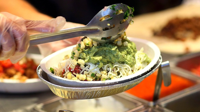 A Chipotle employee adding corn to a burrito bowl with guacamole and shredded cheese