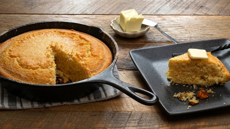 Skillet cornbread next to plated up slice with butter on the side on wooden table.