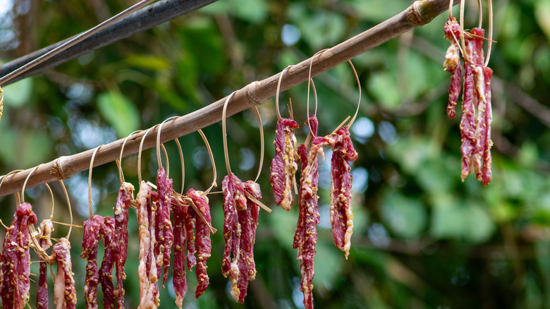 More than a dozen strips of seasoned beef hang from a wooden stick to dry in the sun and become jerky.